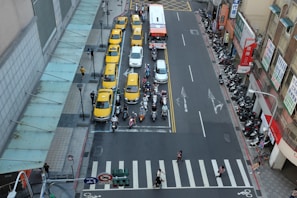 Aerial view of a busy urban street featuring a lineup of yellow taxis and a bus in a dedicated lane. Several scooters are parked and others are waiting at the traffic light. Pedestrians are crossing the street at the crosswalk. The scene is framed by buildings on both sides, with signage visible on one.