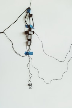An electrical setup with several electronic devices and wires taped to a white wall. Blue tape is used to secure black cables and devices, which appear to be power adapters or similar equipment.
