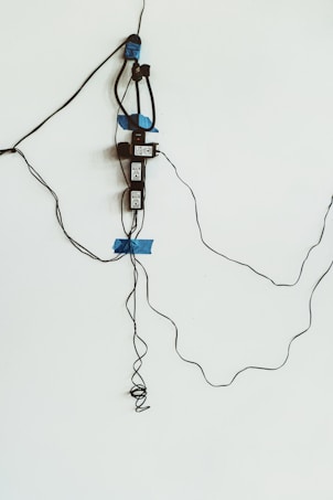 An electrical setup with several electronic devices and wires taped to a white wall. Blue tape is used to secure black cables and devices, which appear to be power adapters or similar equipment.