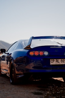 A blue sports car with a spoiler is parked on a sandy terrain. The car has a visible license plate marked with 'Chile'. The background shows a faint outline of a mountain and an expansive pale sky.