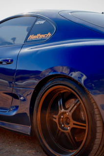 A blue sports car with custom wheels and a visible 'Haltech' sticker on the window. The car is parked on a gravel surface and appears to be in the early evening light, creating a warm glow on the rims.