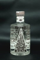 A clear glass bottle filled with liquid, featuring an intricate floral label design in a triangular shape. The label reads Kaja, Special Hand Crafted Batch Distilled Bali Gin, with details of alcohol percentage and volume. The cap is a muted brown color, and the bottle is set against a dark background.