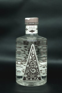 A clear glass bottle filled with liquid, featuring an intricate floral label design in a triangular shape. The label reads Kaja, Special Hand Crafted Batch Distilled Bali Gin, with details of alcohol percentage and volume. The cap is a muted brown color, and the bottle is set against a dark background.