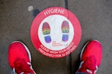 A red circular floor sticker with multicolored shoe prints indicates where to stand. The label emphasizes the importance of hand hygiene, instructing to wash hands frequently for 20 seconds. The sticker is situated on a concrete surface with visible moisture, flanked by a pair of bright red athletic shoes.