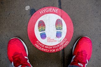 A red circular floor sticker with multicolored shoe prints indicates where to stand. The label emphasizes the importance of hand hygiene, instructing to wash hands frequently for 20 seconds. The sticker is situated on a concrete surface with visible moisture, flanked by a pair of bright red athletic shoes.