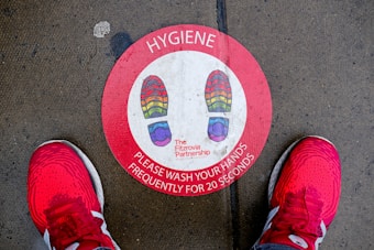 A red circular floor sticker with multicolored shoe prints indicates where to stand. The label emphasizes the importance of hand hygiene, instructing to wash hands frequently for 20 seconds. The sticker is situated on a concrete surface with visible moisture, flanked by a pair of bright red athletic shoes.
