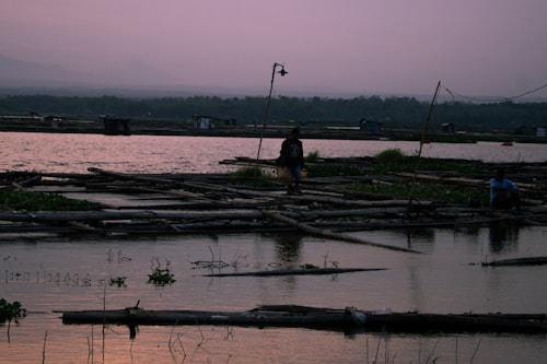 A serene landscape featuring a body of water with wooden platforms and structures floating on the surface. The sky is overcast with a pinkish hue, suggesting dusk or dawn. People are walking and sitting on the platforms, creating a peaceful and quiet setting.