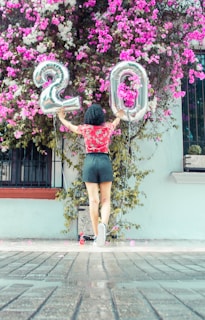 A person stands with their back to the camera, holding large silver balloons shaped like the number 20. The backdrop is a vibrant wall covered with pink and white flowers cascading down, creating a festive and celebratory scene.
