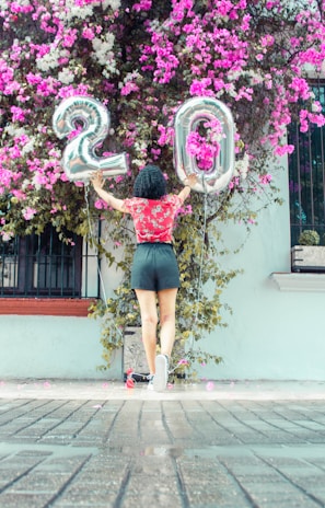 A person stands with their back to the camera, holding large silver balloons shaped like the number 20. The backdrop is a vibrant wall covered with pink and white flowers cascading down, creating a festive and celebratory scene.