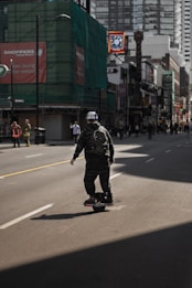 A person wearing dark clothing and a backpack is riding a one-wheeled electric skateboard along a city street. Surrounding the street are tall buildings, some with construction coverings, and various shop signs. The lighting casts shadows across the street, emphasizing the urban setting.