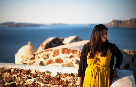 woman in yellow and black jacket standing on rocky ground during daytime in Santorini Greece