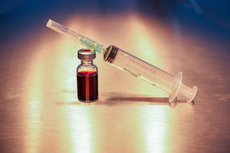 A syringe and a small vial filled with a dark red liquid are placed on a reflective metallic surface. The syringe is labeled and is positioned at an angle, resting against the vial.