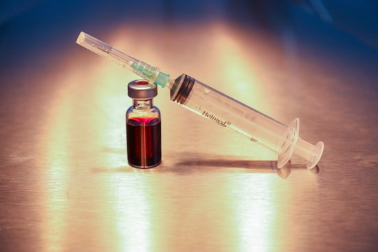 A syringe and a small vial filled with a dark red liquid are placed on a reflective metallic surface. The syringe is labeled and is positioned at an angle, resting against the vial.