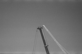 A black and white image of a crane arm extending upward into the sky, with a clear view of the metal structure and cables against a plain background.