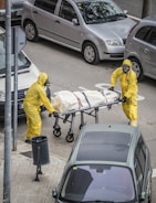 Two individuals in yellow hazmat suits and face masks are handling a stretcher on an urban street. The stretcher appears to have a body bag secured with straps. Several parked cars are visible around them, and there is a trash can on the sidewalk nearby.
