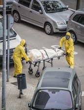 Two individuals in yellow hazmat suits and face masks are handling a stretcher on an urban street. The stretcher appears to have a body bag secured with straps. Several parked cars are visible around them, and there is a trash can on the sidewalk nearby.