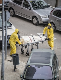 Two individuals in yellow hazmat suits and face masks are handling a stretcher on an urban street. The stretcher appears to have a body bag secured with straps. Several parked cars are visible around them, and there is a trash can on the sidewalk nearby.
