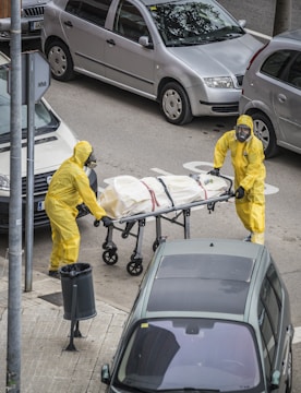 Two individuals in yellow hazmat suits and face masks are handling a stretcher on an urban street. The stretcher appears to have a body bag secured with straps. Several parked cars are visible around them, and there is a trash can on the sidewalk nearby.