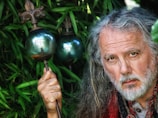 A man with long gray hair and a beard, wearing a red and black patterned shawl, stands among green foliage holding a staff topped with a metallic orb and an ornate fleur-de-lis design. The orb reflects the surrounding nature.