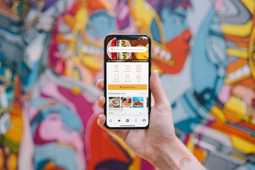 A hand holding a smartphone displaying a user interface for a food delivery app with options for dishes, collections, menus, and places near the user. The background showcases a vibrant and colorful graffiti mural with abstract and humanoid shapes.