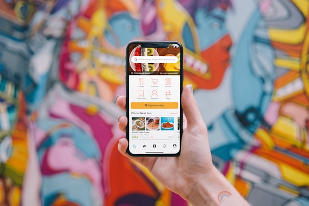 A hand holding a smartphone displaying a user interface for a food delivery app with options for dishes, collections, menus, and places near the user. The background showcases a vibrant and colorful graffiti mural with abstract and humanoid shapes.