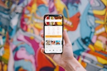 A hand holding a smartphone displaying a user interface for a food delivery app with options for dishes, collections, menus, and places near the user. The background showcases a vibrant and colorful graffiti mural with abstract and humanoid shapes.