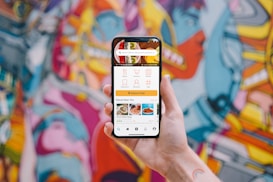 A hand holding a smartphone displaying a user interface for a food delivery app with options for dishes, collections, menus, and places near the user. The background showcases a vibrant and colorful graffiti mural with abstract and humanoid shapes.