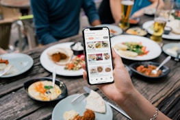 A hand is holding a smartphone displaying a food exploration app at a dining table. The table is set with various plates of food including salads, wraps, and dips. Drinks are also visible, including glasses of beer. Multiple people are seated around the table, visible from the torso down.