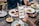 A hand is holding a smartphone displaying a food exploration app at a dining table. The table is set with various plates of food including salads, wraps, and dips. Drinks are also visible, including glasses of beer. Multiple people are seated around the table, visible from the torso down.