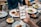 A hand is holding a smartphone displaying a food exploration app at a dining table. The table is set with various plates of food including salads, wraps, and dips. Drinks are also visible, including glasses of beer. Multiple people are seated around the table, visible from the torso down.