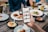 A hand is holding a smartphone displaying a food exploration app at a dining table. The table is set with various plates of food including salads, wraps, and dips. Drinks are also visible, including glasses of beer. Multiple people are seated around the table, visible from the torso down.