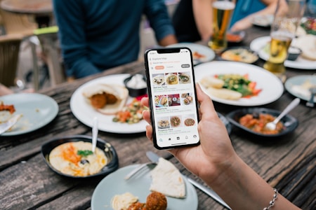 A hand is holding a smartphone displaying a food exploration app at a dining table. The table is set with various plates of food including salads, wraps, and dips. Drinks are also visible, including glasses of beer. Multiple people are seated around the table, visible from the torso down.