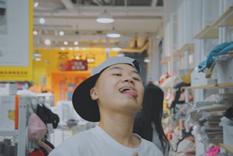 A person wearing a backward cap and a white shirt is playfully sticking out their tongue in a brightly lit store environment. Shelves lined with various items, possibly toys or clothes, are visible in the background. Bright signs with cartoon designs can be seen, contributing to a vibrant and lively atmosphere.