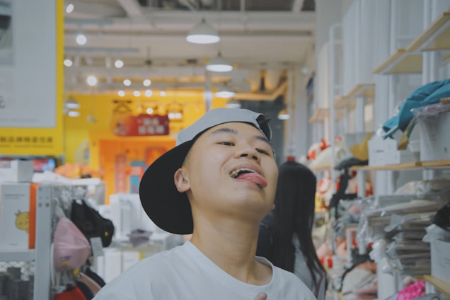 A person wearing a backward cap and a white shirt is playfully sticking out their tongue in a brightly lit store environment. Shelves lined with various items, possibly toys or clothes, are visible in the background. Bright signs with cartoon designs can be seen, contributing to a vibrant and lively atmosphere.