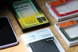 Multiple smartphone cases and packaging boxes are placed on a wooden surface. The cases come in different colors including red, green, and purple, and are neatly encased in their respective packaging. Branding and labels are visible, showcasing details like product names and features.