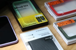 Multiple smartphone cases and packaging boxes are placed on a wooden surface. The cases come in different colors including red, green, and purple, and are neatly encased in their respective packaging. Branding and labels are visible, showcasing details like product names and features.