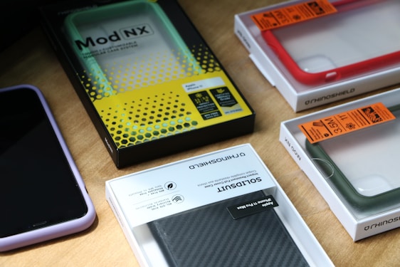 Multiple smartphone cases and packaging boxes are placed on a wooden surface. The cases come in different colors including red, green, and purple, and are neatly encased in their respective packaging. Branding and labels are visible, showcasing details like product names and features.