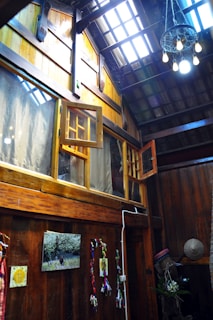 The image depicts a rustic interior space with wooden walls and ceiling. There are large windows, some of which are open, allowing natural light to stream in. A cluster of hanging lights with exposed bulbs adds to the ambiance. Decorations include colorful cloth or ribbons attached to the wall, a framed picture, and a straw hat resting in a corner. Overall, the setting suggests a cozy and traditional atmosphere.