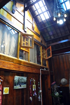 The image depicts a rustic interior space with wooden walls and ceiling. There are large windows, some of which are open, allowing natural light to stream in. A cluster of hanging lights with exposed bulbs adds to the ambiance. Decorations include colorful cloth or ribbons attached to the wall, a framed picture, and a straw hat resting in a corner. Overall, the setting suggests a cozy and traditional atmosphere.