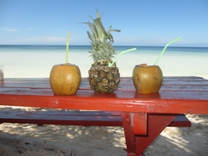 Three tropical fruits placed on a red wooden table on a sandy beach. Two coconuts with straws and a pineapple in the middle are seen with a clear, calm ocean and blue sky in the background.