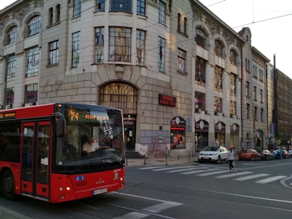 A red city bus is traveling along a street, approaching a crosswalk. The street is lined with a large, historic-style building with multiple windows and a storefront sign reading 'PANDA ROBNA KUCA'. A few people are near the crosswalk, and several parked cars are visible along the street.