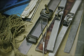 Several metal film splicing tools are arranged on a surface alongside film strips, tangled twine, a metal ruler, and stacks of paper. The scene conveys a vintage or workshop setting, indicative of old-fashioned film editing processes.