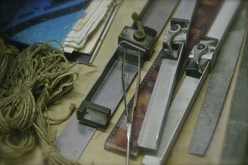 Several metal film splicing tools are arranged on a surface alongside film strips, tangled twine, a metal ruler, and stacks of paper. The scene conveys a vintage or workshop setting, indicative of old-fashioned film editing processes.