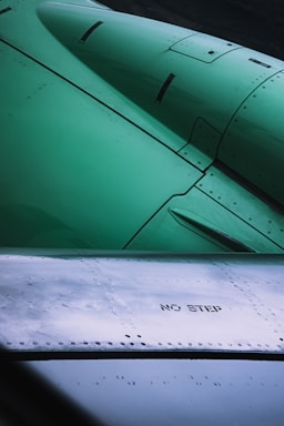 Close-up view of an aircraft wing displaying metal rivets and panels. The text 'NO STEP' is clearly visible on one section of the surface. The upper part of the image shows a green-colored aircraft engine or structure.