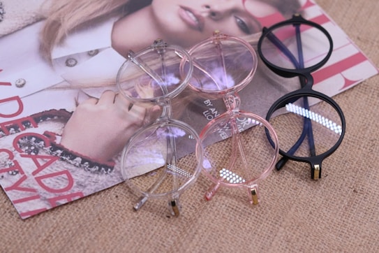 A collection of three eyeglasses with different colored frames - clear, pink, and black - placed on a magazine featuring a fashion photo of a woman. The background is a textured burlap surface.
