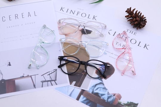 Four pairs of eyeglasses are laid out on top of lifestyle magazines. The glasses have different frame colors: black, pink, blue, and clear. A pinecone lies nearby, adding a decorative element to the setting. The magazines display minimalist designs and text, contributing to a clean aesthetic.