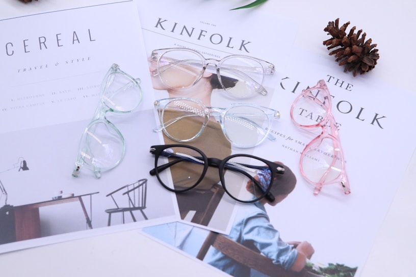 Four pairs of eyeglasses are laid out on top of lifestyle magazines. The glasses have different frame colors: black, pink, blue, and clear. A pinecone lies nearby, adding a decorative element to the setting. The magazines display minimalist designs and text, contributing to a clean aesthetic.