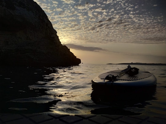 A paddle board rests on gentle, rippling water as the sun sets, casting a golden hue over the scene. The sky is filled with scattered clouds, and a large rock formation rises on the left side of the image.