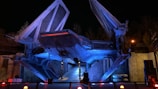 A large, futuristic spacecraft illuminated by blue and red lights stands against a night sky. The vessel has angular wings swept upwards and intricate mechanical details. Surrounding the ship are industrial elements, including metal structures and pipes.
