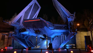 A towering alien woman in 1950s sci-fi style stands beside a sleek spaceship under a twilight sky